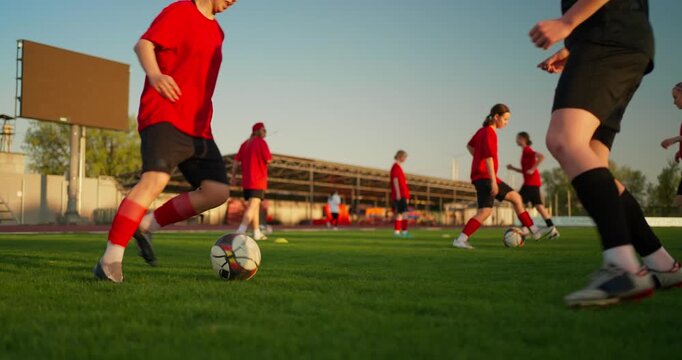 Young female soccer players in red uniforms dribble and train with balls on a green outdoor field during warm evening practice