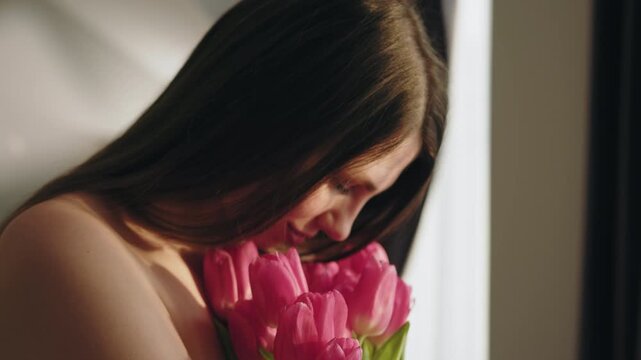 Young woman with long brown hair gently smelling a bouquet of vibrant pink tulips indoors with soft natural light. The serene moment conveys tenderness, joy and the delicate beauty of floral fragrance