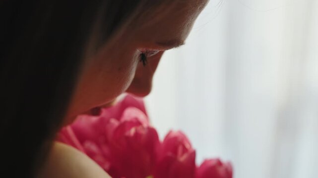 Close-up profile of young woman inhaling fragrance from vibrant pink tulips against soft blurred background, conveying peaceful moment. The delicate detail highlights tenderness, joy and the graceful
