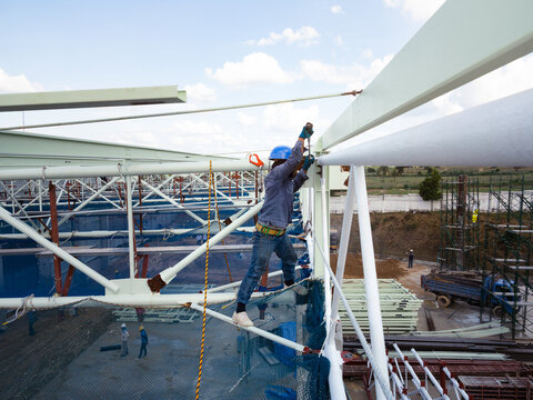 Construction worker wearing blue safety helmet and protective harness uses torque wrench to tighten nuts and bolts while assembling roof truss steel structure. Industrial construction scene.