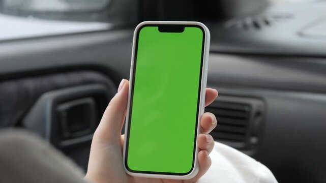 Hand holds phone with green screen inside car. Person sits relaxed, focused on device. Useful for driving, navigation, or app promotion. Green screen allows easy overlay of content