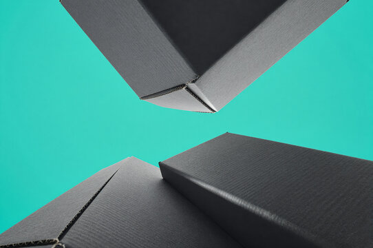 Cardboard box components hover in an asymmetrical arrangement against a solid teal backdrop. Dark gray matte surfaces show fine paper texture under even studio illumination.