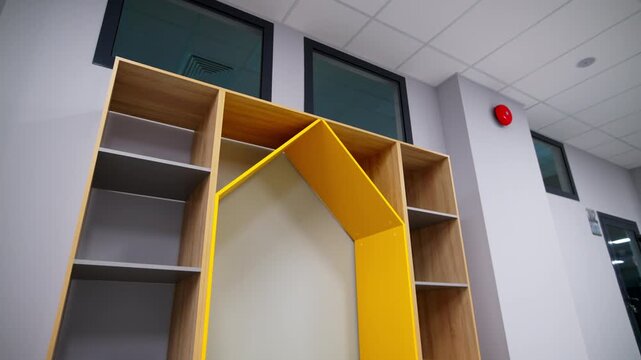Wooden cabinet in house shape stands in spacious classroom. Colorful shelves and small roof create playful furniture piece for children