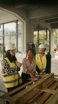 Three professional women in hard hats and safety vests discuss blueprints at a construction site. A diverse team of female architects and engineers collaborate on project plans.