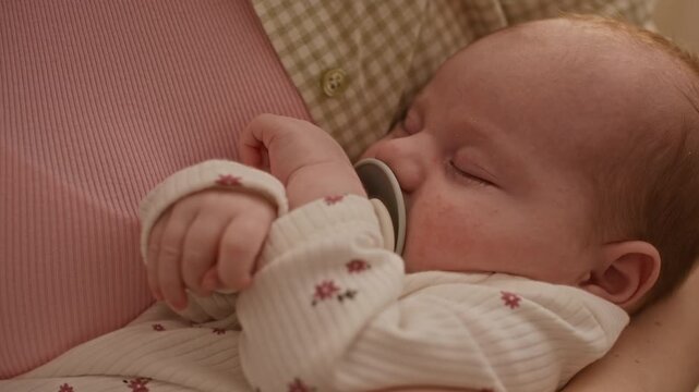 Closeup shot of Caucasian infant girl in soft white onesie with floral pattern sleeping in caring arms of her unrecognizable mother with pacifier in mouth
