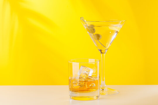 Glass of whiskey with ice and martini with olives on yellow background