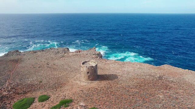 Aerial view circling the ancient stone watchtower of Torre di Cala Domestica on a rocky cliff overlooking the beautiful blue sea in Sardinia, Italy