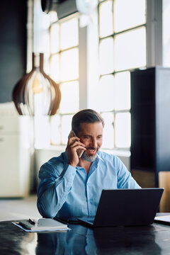 Happy CEO working on laptop while talking on cell phone in office.