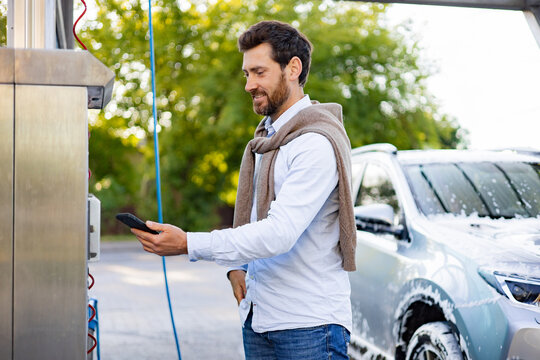 A smiling Caucasian man uses his smartphone to pay for a car wash service at an outdoor automated station