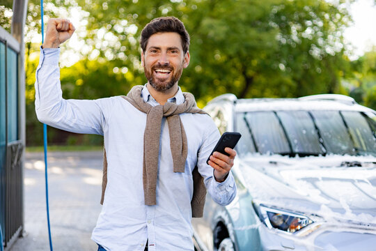 A happy caucasian man holds his phone and raises his fist in triumph while standing next to his freshly washed, foamy car