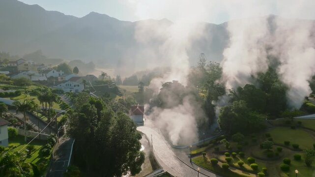 Aerial view of the village of Furnas on Sao Miguel Island, showcasing volcanic steam vents alongside a winding stone road, manicured gardens, and lush surroundings bathed in soft sunlight.