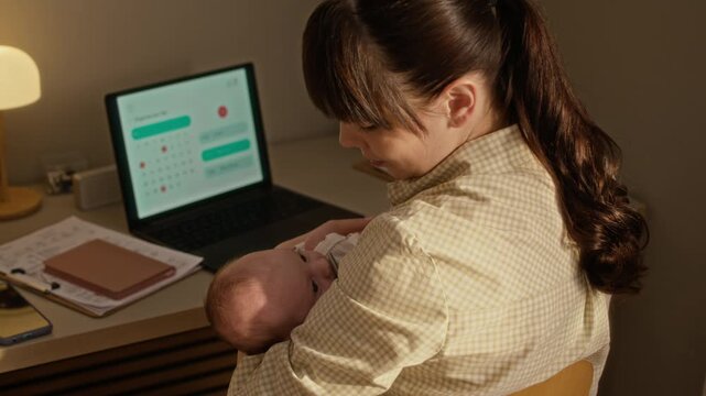 Arc shot of young Caucasian mother rocking infant baby girl in arms while sitting at table in front of laptop, working from home in evening