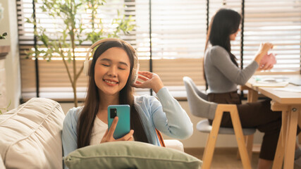 Fototapeta premium Happy Young Asian Woman Enjoying Music with Wireless Headphones and Smartphone, Smiling and Relaxing on Cozy Sofa in Modern Living Room.