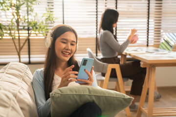 Fototapeta premium Happy Young Asian Woman Enjoying Music with Wireless Headphones and Smartphone, Smiling and Relaxing on Cozy Sofa in Modern Living Room.
