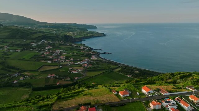 Aerial view of a charming coastal village on Faial Island, Azores, featuring white houses with orange roofs set amidst vibrant green fields, overlooking the Atlantic Ocean and a volcanic coastline.