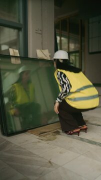 A professional woman in a hard hat and safety vest applies makeup using the reflection in a large glass panel at a construction site. She takes a moment for self-care before continuing her workday.
