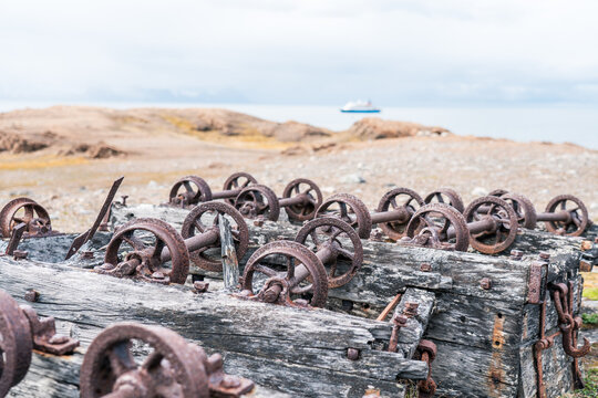 Svalbard, Svalbard and Jan Mayen - 05 July 2025: View of rusted iron wheels and weathered wooden remains at Varsolbukta with a ship in the distance under an overcast sky.