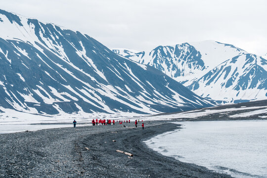 Svalbard and Jan Mayen - 02 July 2025: View of a group of tourists in red jackets standing on a dark pebble beach with snow-capped mountains and calm arctic waters.