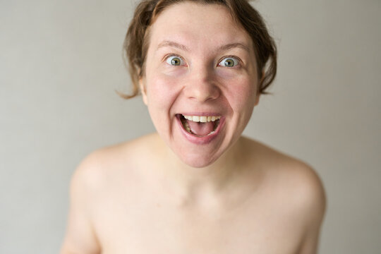Young Woman Shouting With Wide Open Mouth In Neutral Indoor Background Bare Shoulders Visible Close-Up Portrait Showing Energetic Surprised Facial Expression.