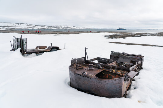Svalbard, Svalbard and Jan Mayen - 21 June 2025: View of abandoned rusty vehicles at Kinnvika Station surrounded by snow with a ship and tourists in the distance.