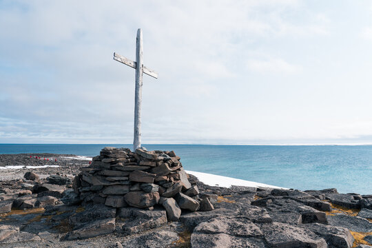 Svalbard, Svalbard and Jan Mayen - 24 April 2026: View of a weathered wooden cross standing on a stone cairn along the rocky coastline with the Arctic Ocean and patches of snow under a bright sky.