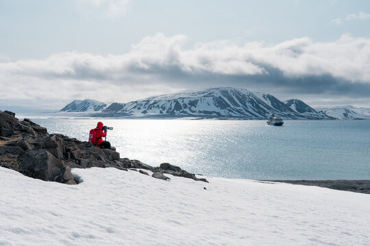 Svalbard, Svalbard and Jan Mayen - 24 April 2026: View of a photographer in a red jacket sitting on a rocky outcrop overlooking a cruise ship in arctic waters surrounded by snow-capped mountains.