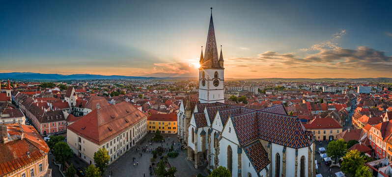 Aerial panorama of the old town of Sibiu featuring historic red roofs and the tall church tower, with the majestic blue Fagaras mountains visible on the horizon. Romania Transylvania, landscape.