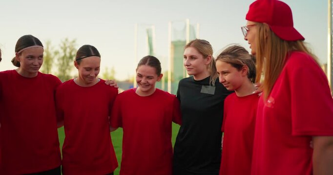Youth girls soccer team in red jerseys gathers in a supportive huddle with coach on outdoor field before practice at sunset