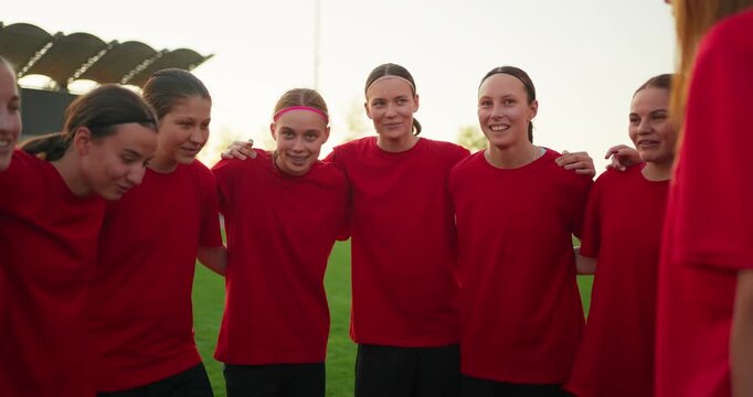 Youth girls soccer team in red jerseys standing arm in arm during a supportive pregame huddle on a sunny outdoor field with stadium seating