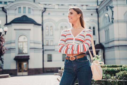 Young woman smiling confidently in a casual striped sweater and jeans standing in a sunlit city street with a shoulder bag