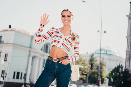 Smiling young woman waving on a sunny city street wearing striped crop top and jeans, casual summer fashion and carefree urban lifestyle