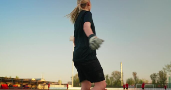 Female soccer goalkeeper standing on outdoor stadium field at dusk, holding a ball and watching teammates during practice from behind
