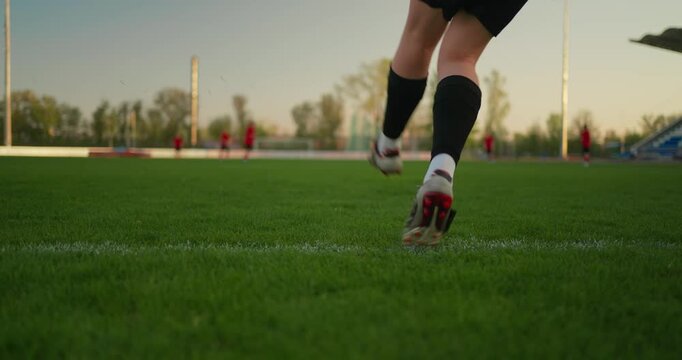 Low-angle view of a soccer ball resting on green field turf as a player approaches, with blurred teammates, goalposts, trees, and warm evening light