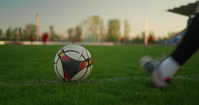 Close-up soccer ball on a green field touchline at sunset, with players training in the background and a stadium stand beside the pitch