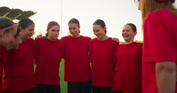 Teenage girls soccer team in red jerseys standing arm in arm during a pregame huddle on a grassy field, smiling with unity and focus