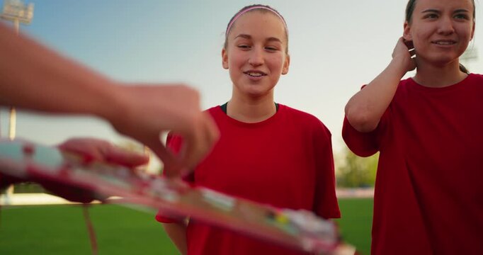 Teenage soccer players in red jerseys gather on a field as a coach points to a tactics board during an outdoor team strategy session