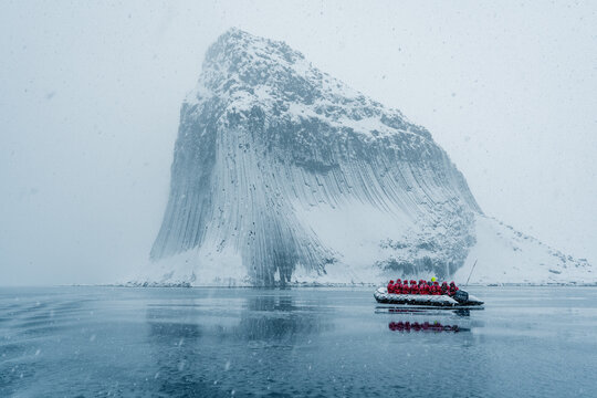 View of the snow-covered cliffs of Antarctica with vertical columns and a boat full of people in red parkas during a snowfall in Antarctica.