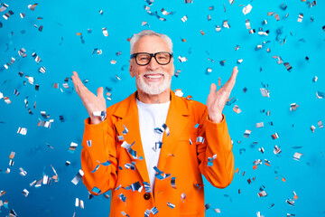 Obraz premium Joyful senior man wearing orange jacket and glasses celebrating with silver confetti shower on bright blue background, smiling and raising hands in festive party mood