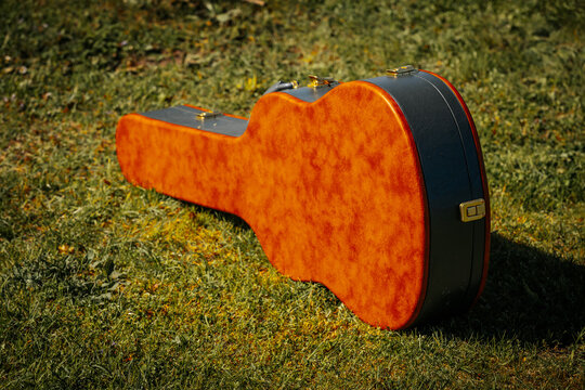 Acoustic guitar case with a vibrant orange finish resting on green grass under natural sunlight in an outdoor setting