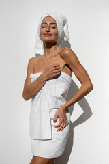 Woman wrapped in white towel with hair towel standing against white wall, enjoying spa wellness moment with closed eyes, natural light, minimal self care concept and relaxation © sutulastock