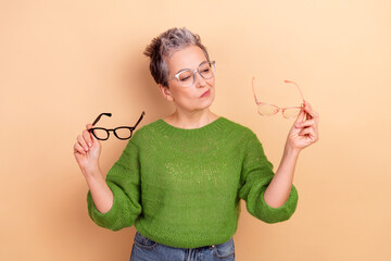 Fototapeta premium A confident mature lady in a green sweater holds two pairs of glasses against a beige background showcasing style fashion and a playful thoughtful expression
