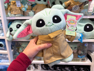 Naklejka premium Dubai, UAE - 24 may 2026: Hand holding Grogu plush character in modern toy store surrounded by collectible Star Wars merchandise, representing entertainment retail and fandom culture lifestyle