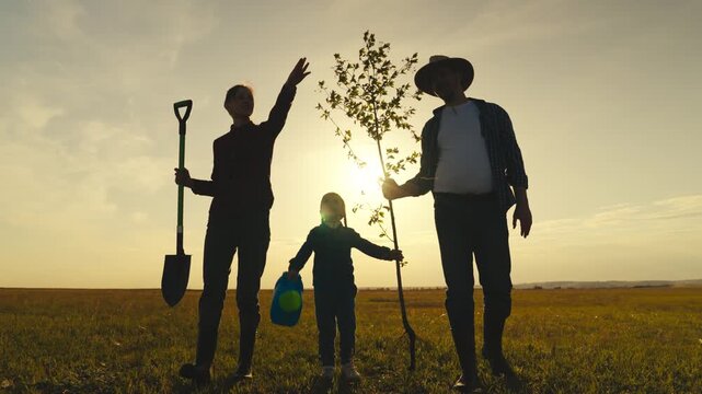 A family of three, a man and two children, are planting a tree in a field. The man is wearing a hat and holding a shovel, while the children are holding a tree sapling
