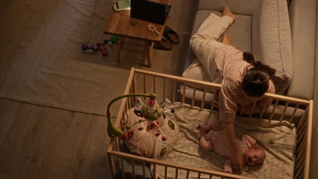 High angle full shot of loving young Caucasian mother lying on couch near wooden crib in nursery room and comforting crying newborn baby at night