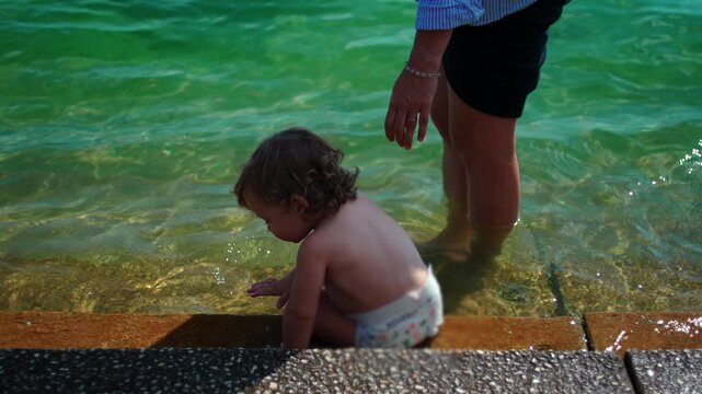 Toddler sitting at edge of shallow water beside parent during summer outing capturing curiosity, sensory exploration and authentic outdoor childhood experience