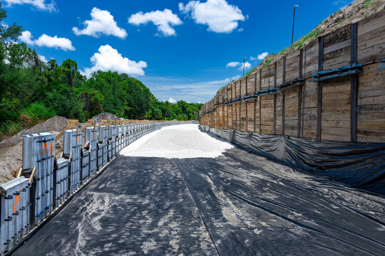 Perspective view of an active civil engineering construction site showing a newly prepared roadbed lined with black geotextile fabric and white gravel, flanked by a tall wooden shoring wall and concre