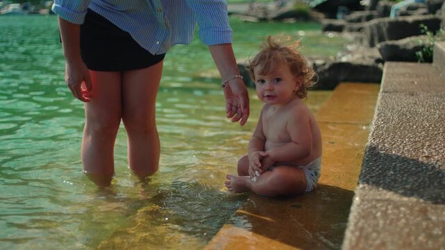 Toddler sitting at shallow lakeside edge beside mother during summer outing capturing sensory exploration, curiosity and authentic outdoor childhood freedom