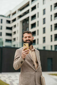 Smiling bearded businessman using smartphone standing in urban setting