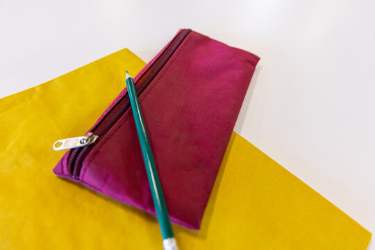 Magenta zippered pouch lying on white desk with sharpened green pencil, yellow paper, metal pull
