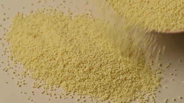 Close-up shot of dry organic couscous grains pouring from a bowl onto a pile on a beige background. Healthy Mediterranean food ingredient. 
